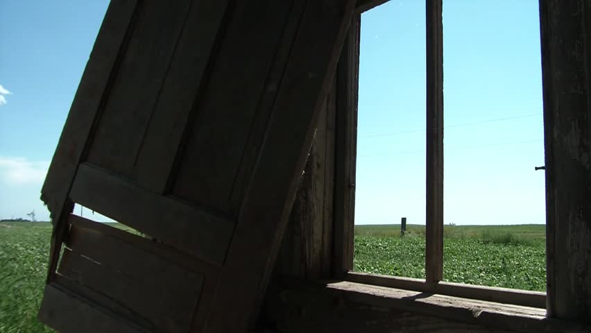 Man pretends to knock on window without glass at old abandoned farm house with lush green field in rural background.