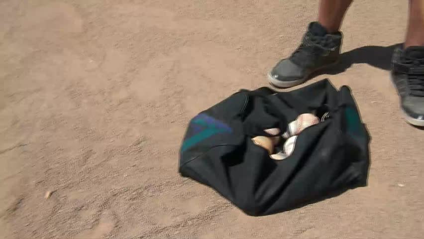 Pan from a bag of balls to a baseball bat being tossed onto home plate in sunlight.