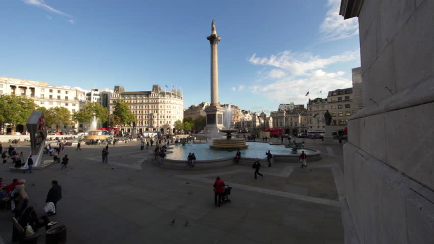 A panoramic shot of Trafalgar Square in London. Lord Nelson's monument, the Olympic clock, buildings and people can be seen. 