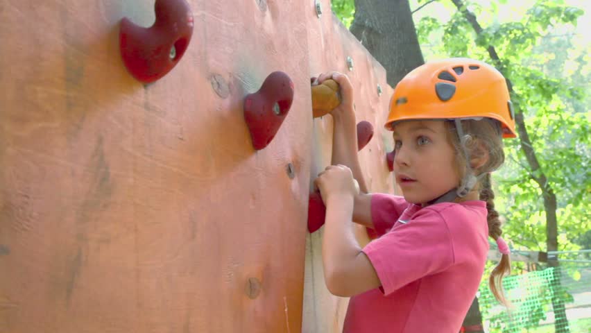 Rock climbing. Little girl in helmet crawls on pendant wall in outdoor climbing center, closeup
