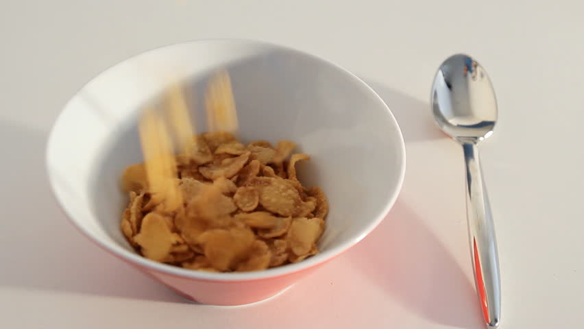pouring flakes into a breakfast bowl with a spoon beside the bowl on a white table