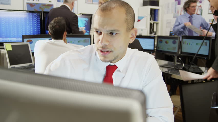 Young and ambitious stock market trader is doing a deal over the phone in a busy office filled with computers. The rest of his team are hard at work in the background. 