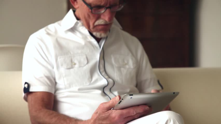 Senior man using tablet sitting on white couch in living room. Starting and ending blurred. (No audio).