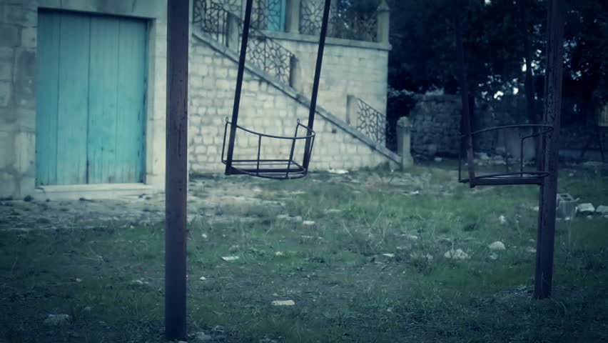 A swing is moving in a backyard, but no one is there. Horror themed shot, medium shot.