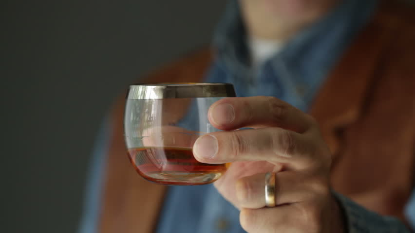Whisky drinker wearing a denim shirt and leather vest presents a glass of whisky to the camera with his left hand. Recorded with macro lens and shallow depth of field.
