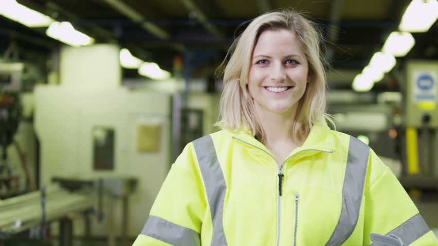 Portrait of a cheerful and friendly female warehouse manager who is wearing a hard hat and reflective clothing. She takes off her hard hat and smiles at the camera.  In slow motion.