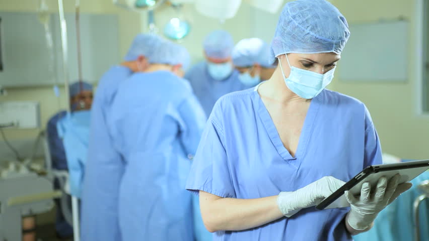 Female Caucasian trauma nurse using wireless tablet while skilled medical professionals perform surgery operating theater
