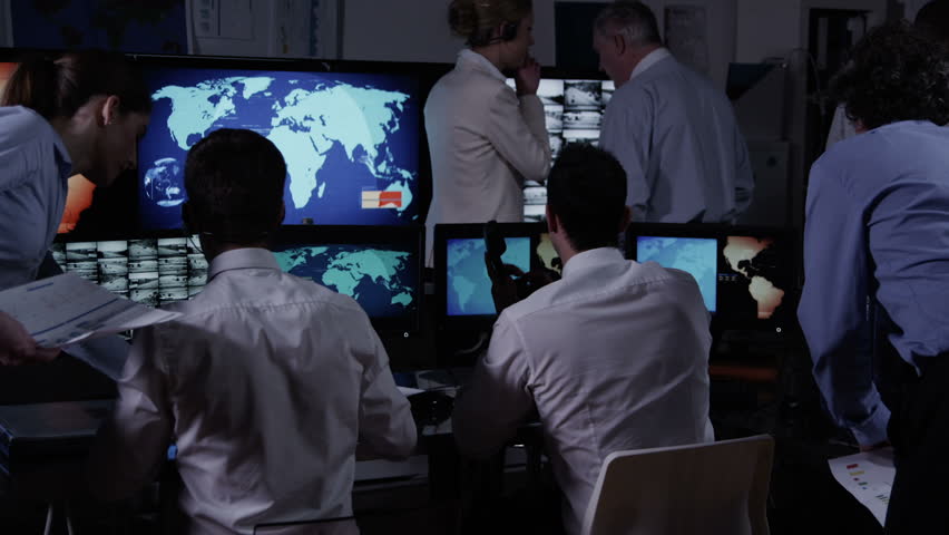 A team of security personnel working in a busy system control room, could be a weather station/airport traffic control. It could be a  power station or police/army control facility. In slow motion.