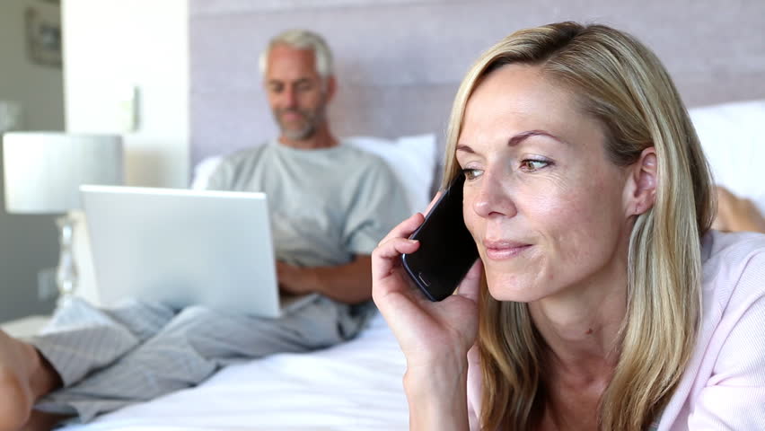 Woman laughing while she is on the phone next to his husband on his laptop in the bed