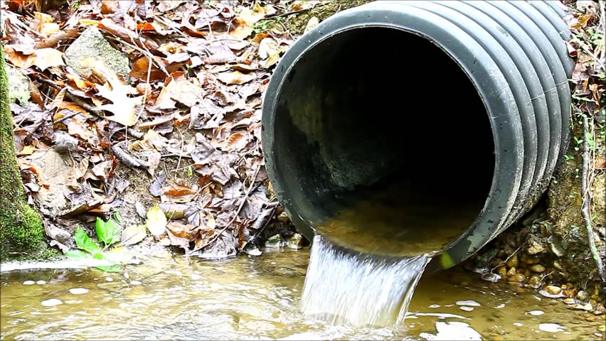 A black plastic storm drain pipe with storm water runoff flowing out of it into a spillway leaching into the local water table 
