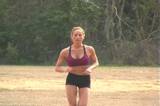 A beautiful young woman jogs toward the camera in a grassy field.  Camera zooms out and pans left to follow, allowing the woman to jog out of frame left.