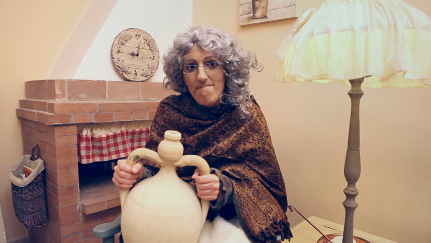 Funny scene of a granny (a dressed up actress) with a scarf, on a rocking chair, eagerly drinking some alcoholic liquid from an earthenware jug. Humorous shot.