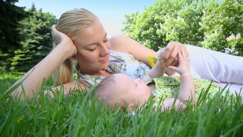 Young woman lies near her baby daughter and they play with yellow dandelion, on grass at sunny summer day in park