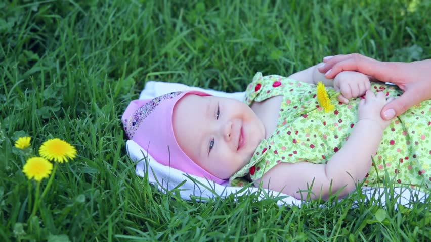 Baby girl lies on grass and hold dandelion, mother hand touches her at sunny summer day