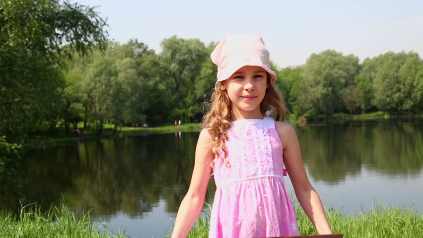 Little girl raises picture frame and smiles near pond at summer park