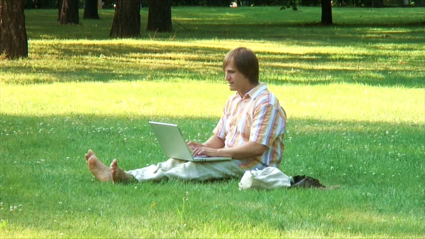 Barefoot man types on laptop siting on a grass in a park