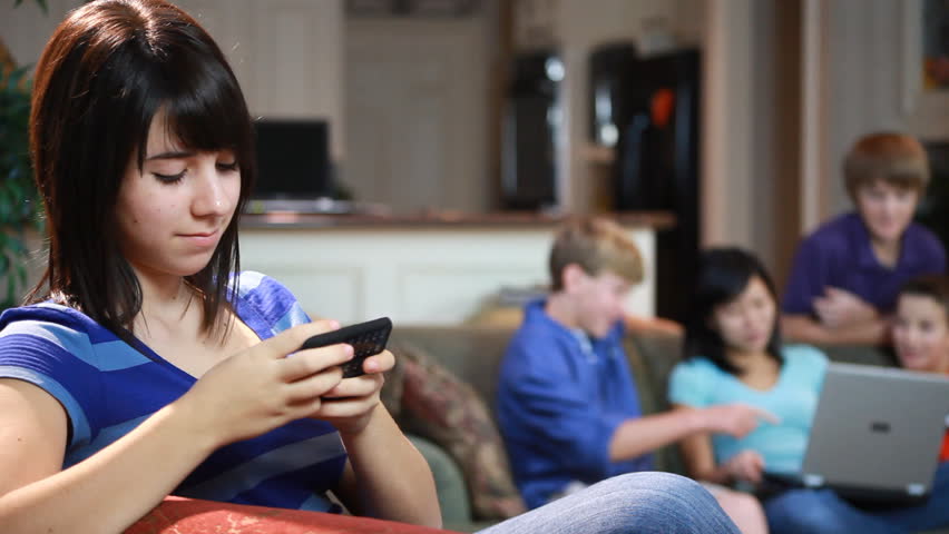 A teenage girl texting on a cell phone to an out of focus group of students on the couch with a laptop.