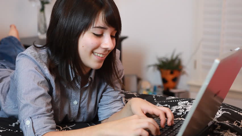 Two teenage girls lying on a bed, one using a laptop and the other text messaging on a cell phone. (camera pan from one girl to the other)