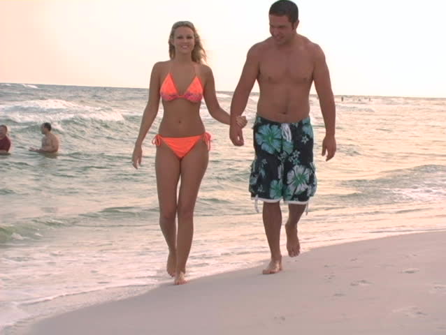 couple walking in edge of water - toward camera - tilting up.  Woman in orange bikini