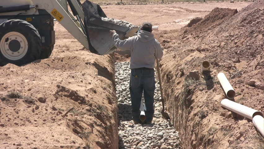 A man gives hand signals to the backhoe operator who is dumping rocks into a new drain field.