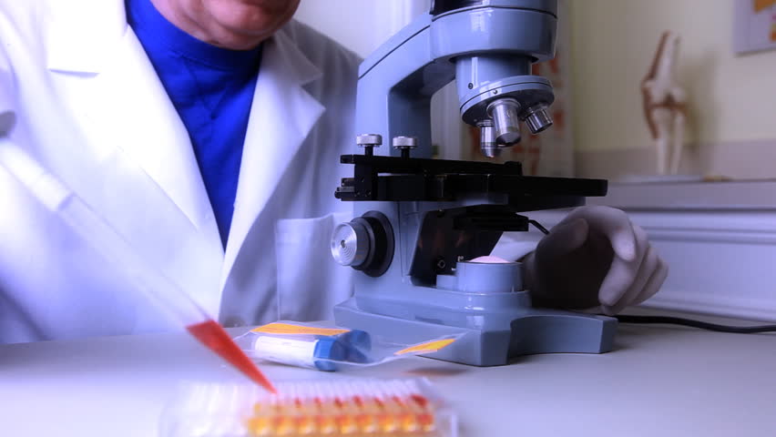 A biologist, scientist, or lab tech places a sample under the microscope with a pipette, then examines the specimen. Tinted for affect.