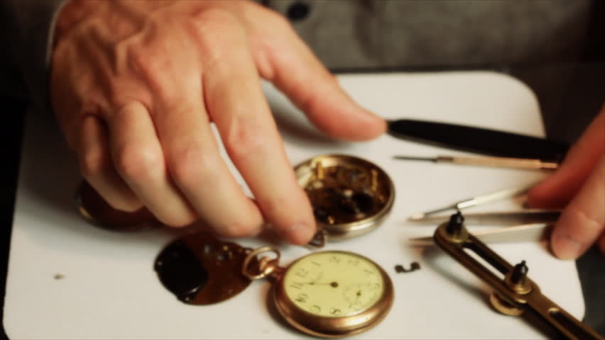 A man wearing jeweler's glasses, picks up an old pocket watch and examines the inside mechanisms.