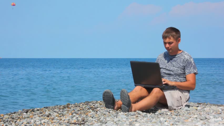 man with notebook sitting on pebble beach, parasailing on sea in background 