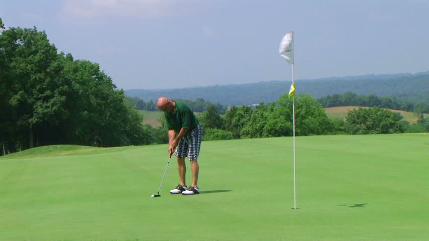 Golfer uses putter to sink long putt into hole with flagstick, on country club course. 