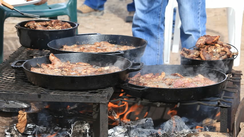 Lamb steaks and chops cooking in caste iron pans over an open outdoor fire. Men turning meat.  Cowboy and sheep camp bbq and party. Fresh meat with smoke and oil boiling near Dutch Oven. 