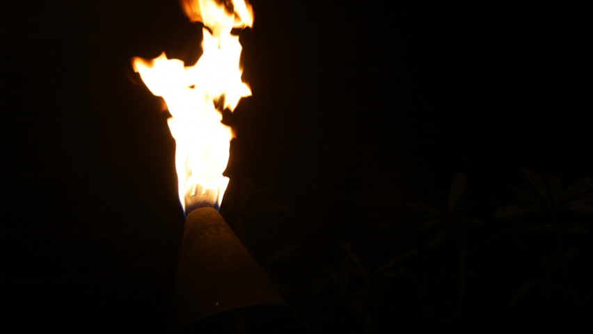 A tiki torch flame blowing in the wind at night on Kauai, Hawaii. Palm trees are faintly visible in background at times.