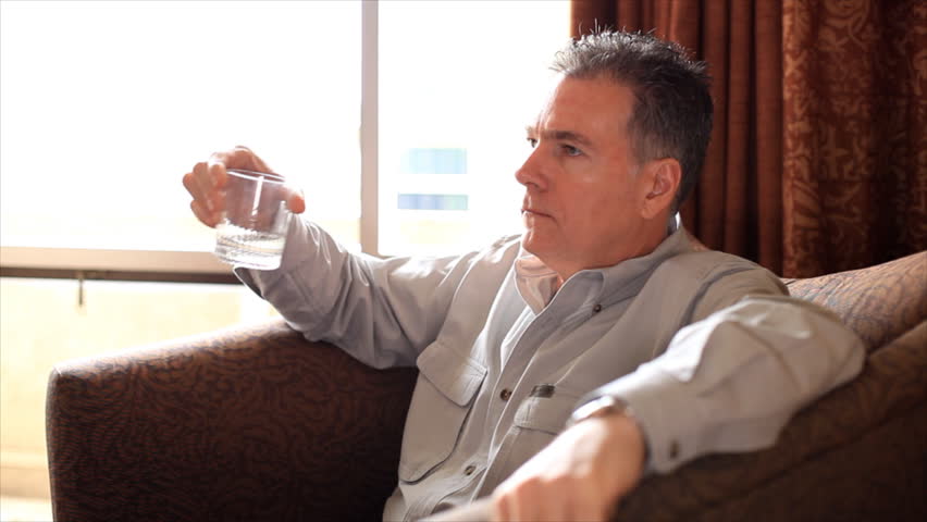 A pensive man sitting by a big bright window in a large lounge chair drinking something from a clear glass.