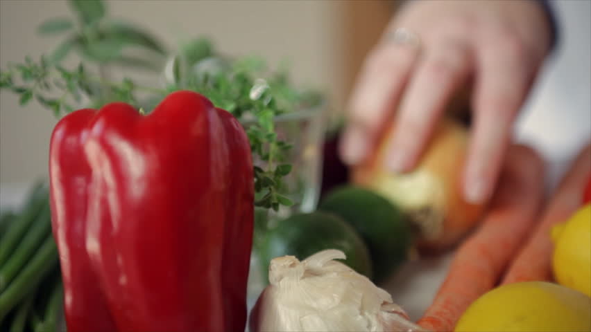 The camera pans across a variety of vegetables to focus on a woman peeling a flavorful yellow onion. Pan, Tilt, Rack focus.