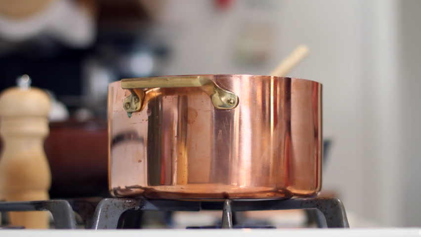 A woman chef stirs green beans into a gleaming copper pot she is using to cook on her gas stove and then she adds pepper from a pepper mill for seasoning.