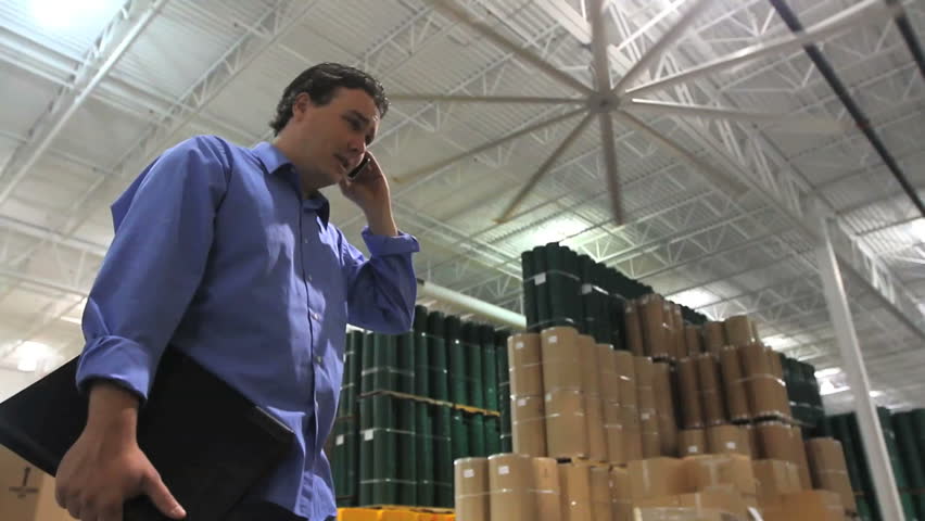 A supervisor talks on his phone in the middle of a large industrial warehouse. 
